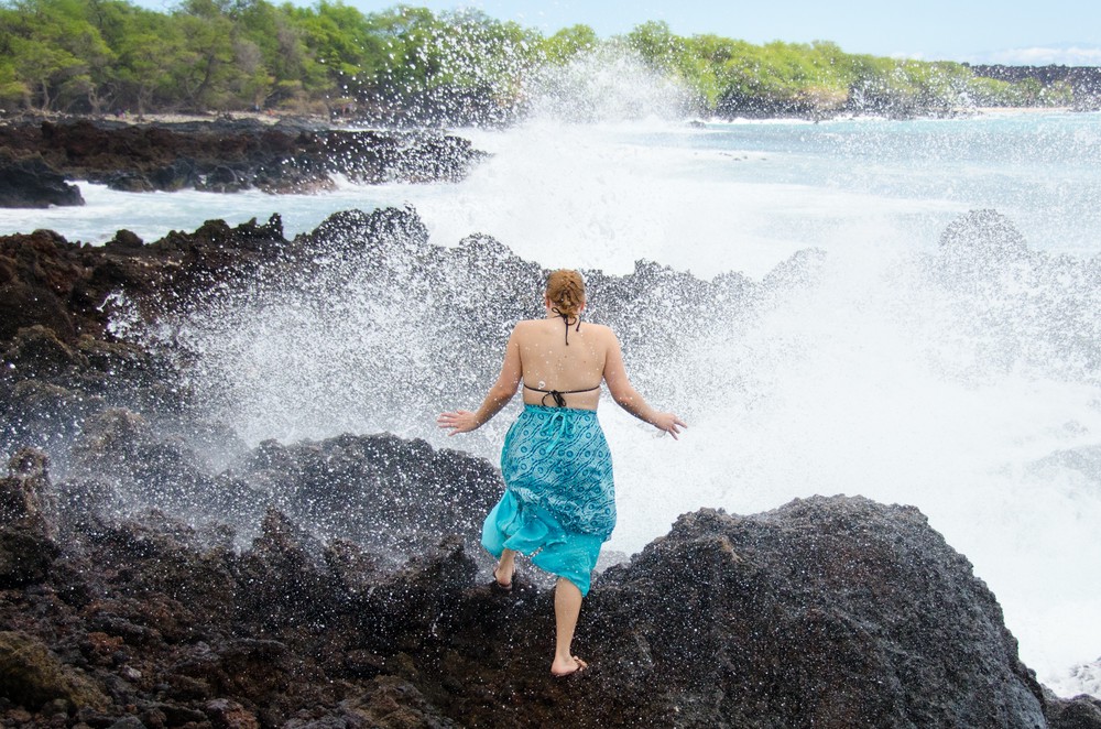 A woman stands on a rock looking away wearing a swimsuit and turquoise dress. A wave has just crashed and sprayed the woman with water surprising her. She holds her hands out to the side.
