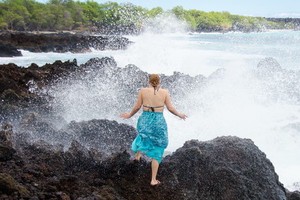 A woman stands on a rock looking away wearing a swimsuit and turquoise dress. A wave has just crashed and sprayed the woman with water surprising her. She holds her hands out to the side.