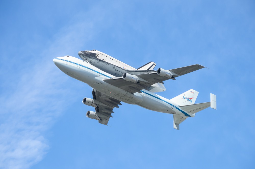 In the blue sky a jumbo jet flies with a space shuttle attached to its back.