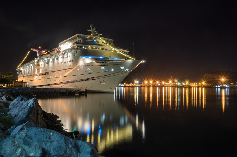 A large cruise ship is lit by lights on the left side of the image. To the right are the lights of a dock reflected in the water of the harbour.