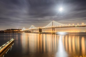 At night a suspension brdige is seen spanning a river from left to right. The bridge is lit up by lights on the cables. There is a pier in the bottom left and the moon is in the sky in the top right.