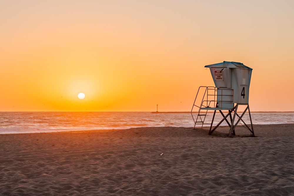 A beach at sunset, the sun is just above the horizon still. A lifeguard station stands to the right side of the image.
