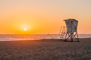 A beach at sunset, the sun is just above the horizon still. A lifeguard station stands to the right side of the image.