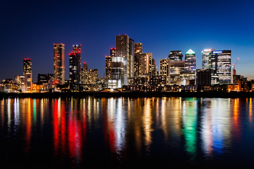 The tall buildings of London shot over the River Thames at night. The buildings arte lit with lights which reflect off the smooth surface of the river.